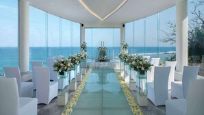 An indoor ceremony space with a glass floor, white chairs, and floral arrangements, overlooking a coastal view. - Hilton Bali Resort (Nusa Dua)