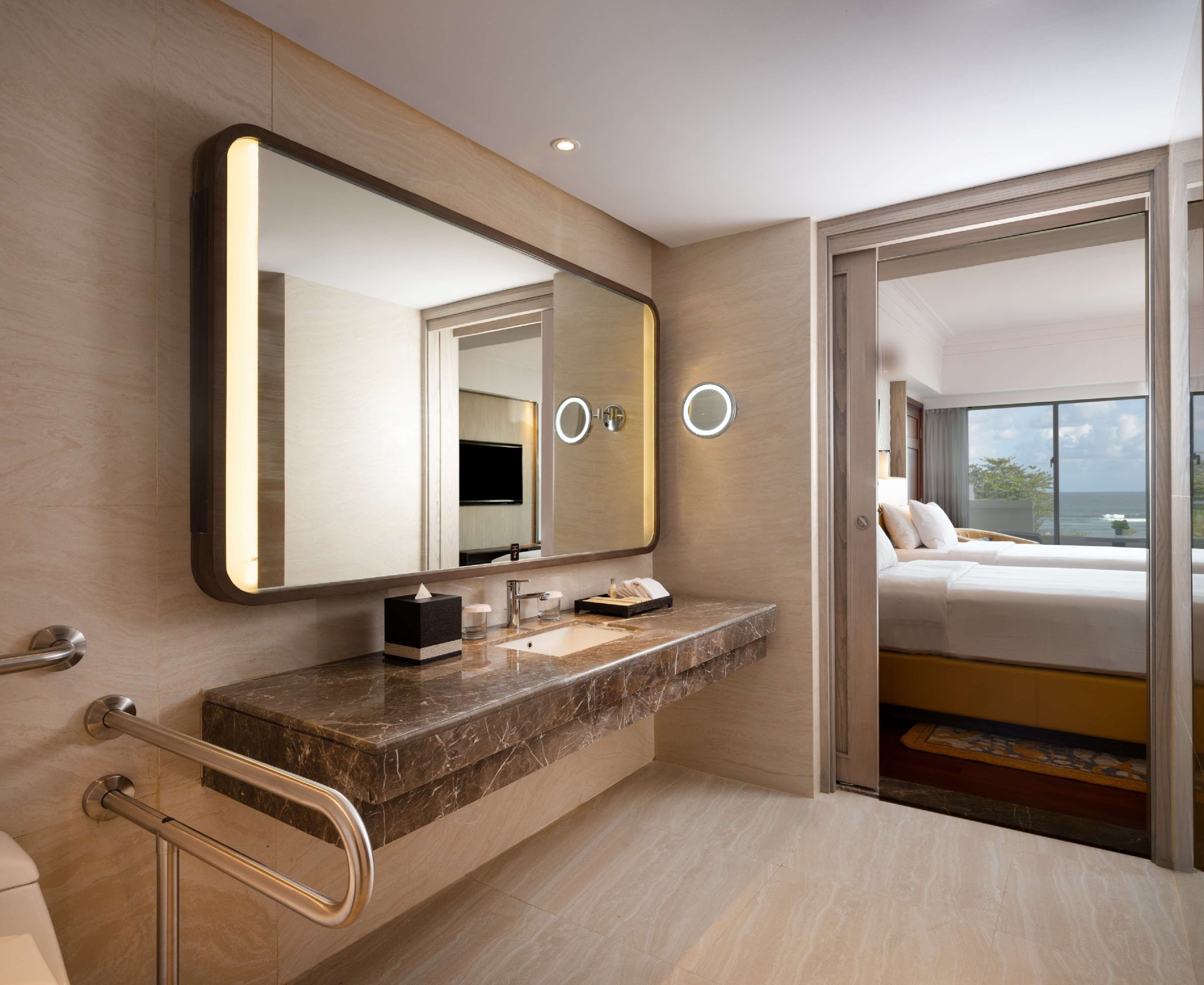 Een moderne badkamer met een marmeren aanrecht, een grote spiegel en een zichtbaar bed met uitzicht op de oceaan.