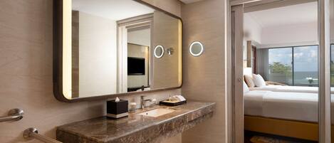 A modern bathroom with a marble countertop, a large mirror, and a visible bed with a view of the ocean.