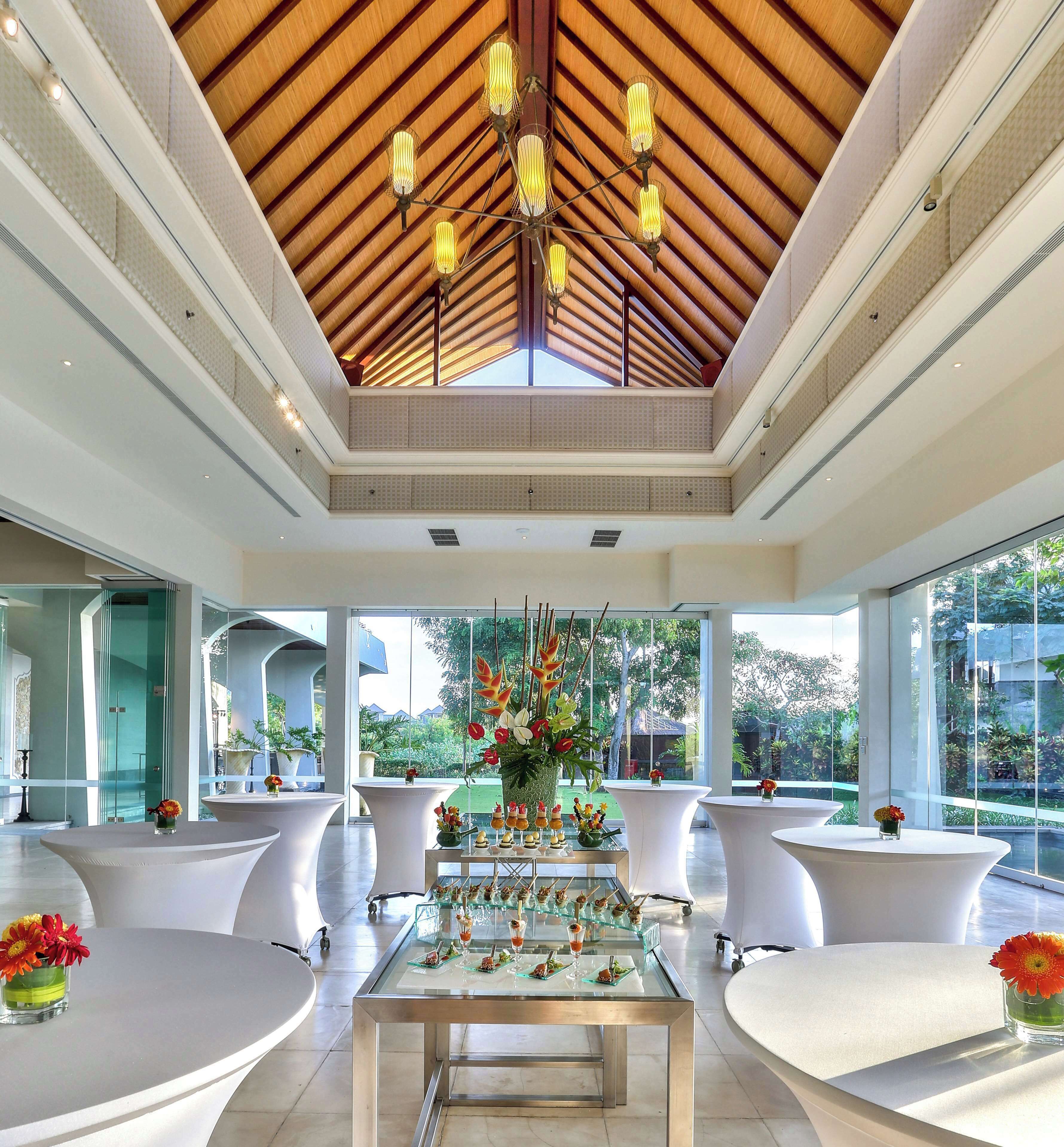 A modern hotel lobby with a high ceiling, large windows, and a central glass table displaying appetizers.