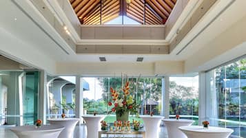 A modern hotel lobby with a high ceiling, large windows, and a central glass table displaying appetizers.