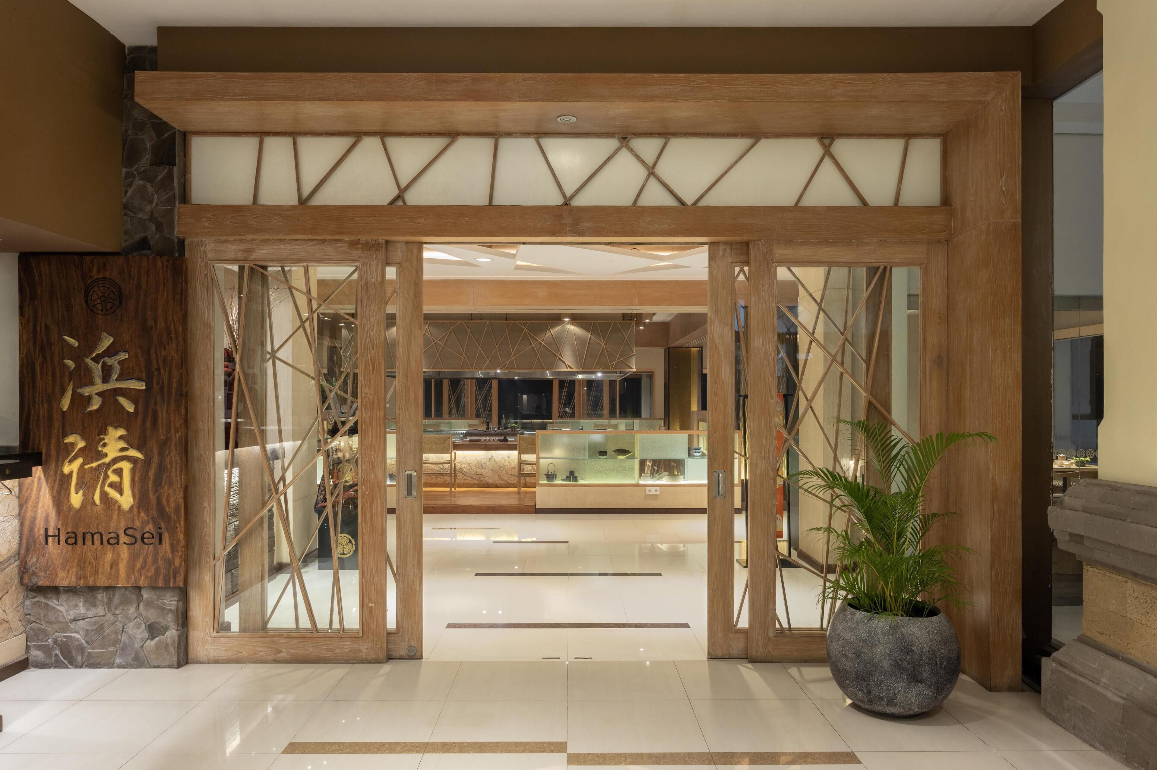 A hotel entrance with a wooden and glass facade, a reception desk, and a potted plant.