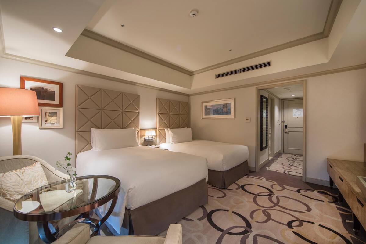 A hotel room with two beds, a glass table, a chair, a lamp, and a patterned carpet.