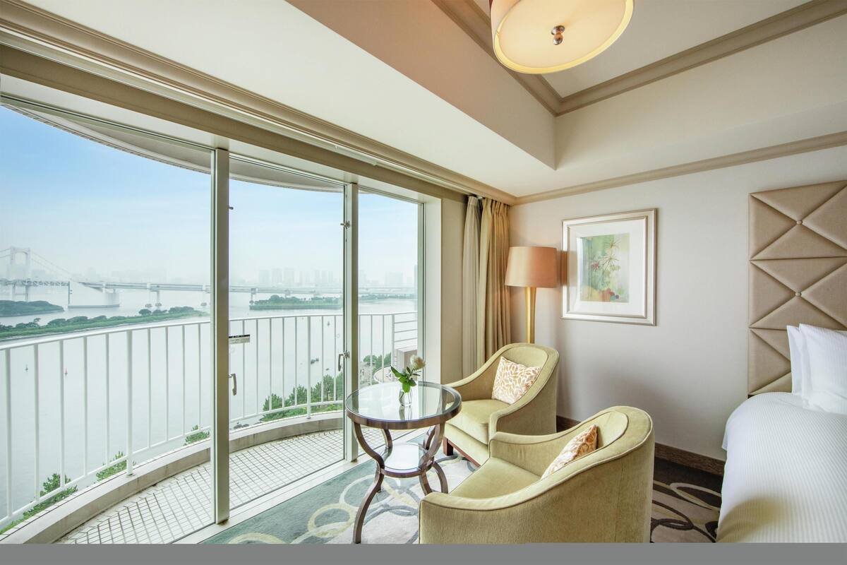 A hotel room with a large window, a bed, a round table with a vase, two armchairs, a lamp, and a framed picture on the wall.