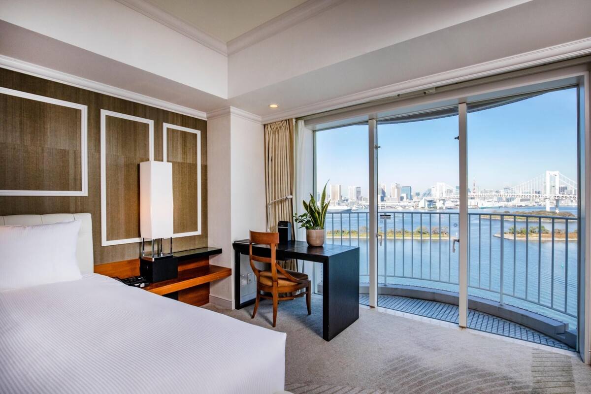 A hotel room with a bed, desk, chair, potted plant, and a large sliding glass door leading to a balcony with a city view.