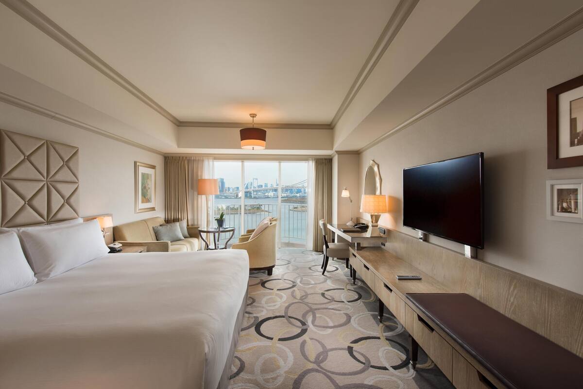 A hotel room with a large bed, a desk with a chair, a TV, and a view of a cityscape through the window.