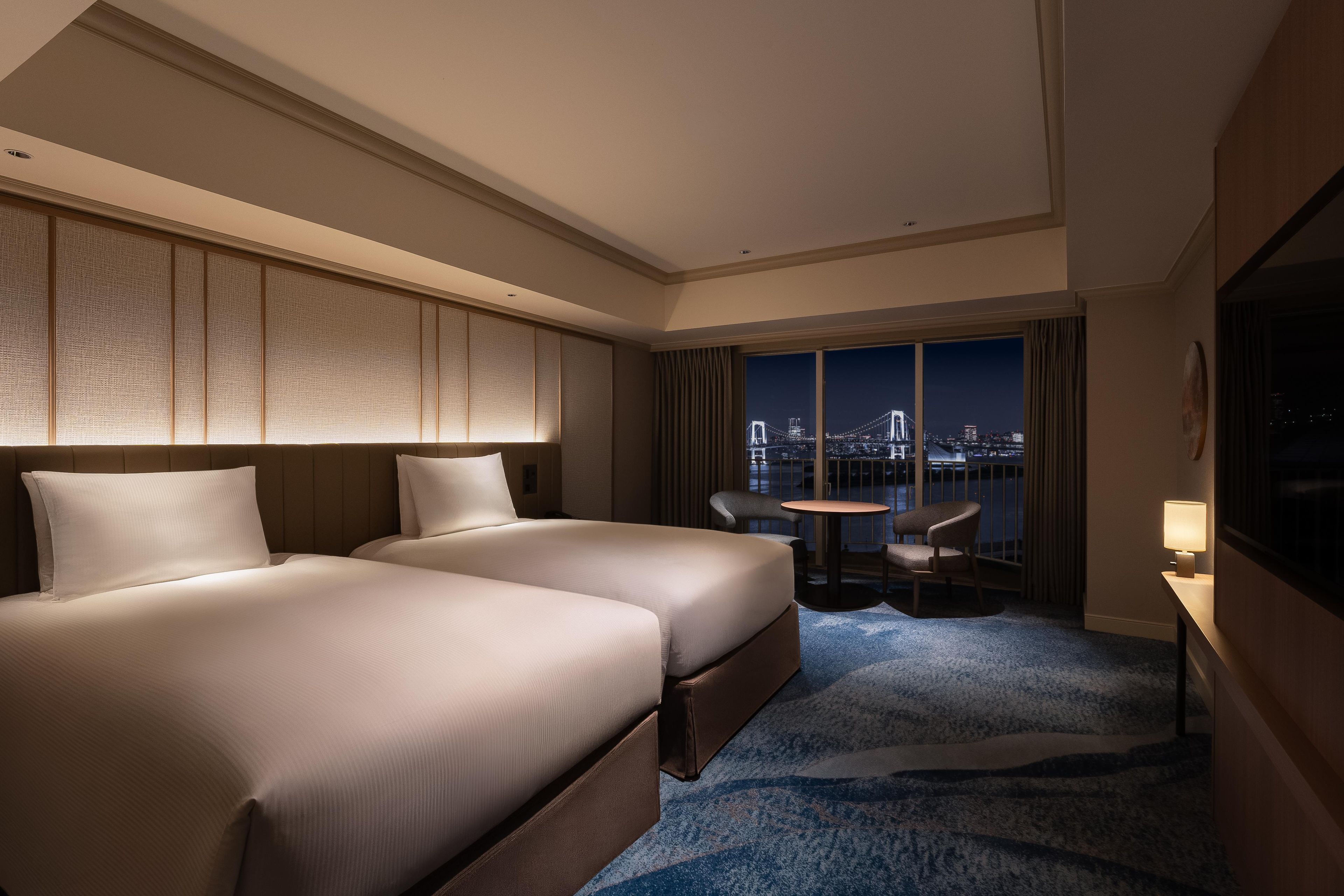 A hotel room with two beds, a view of a cityscape, a small table, and a chair.