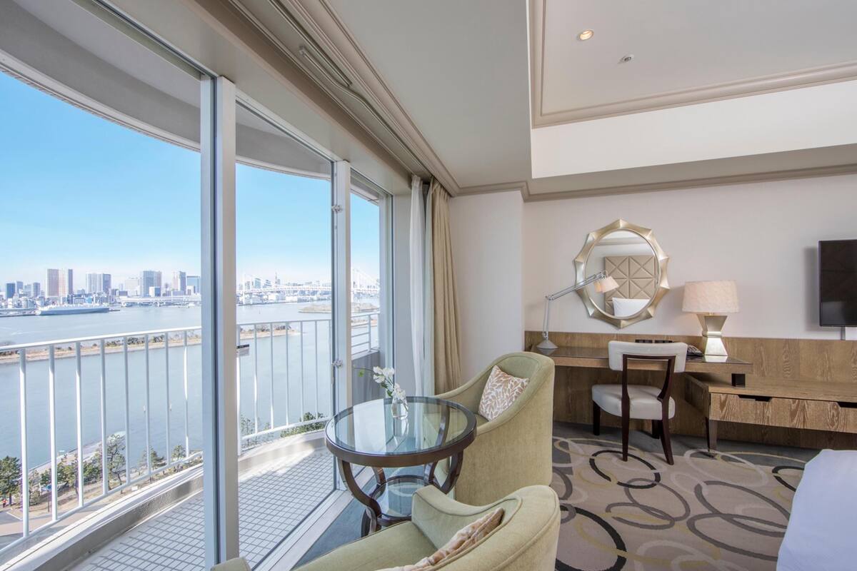 A hotel room with a balcony, a desk, a chair, a bed, and a TV.