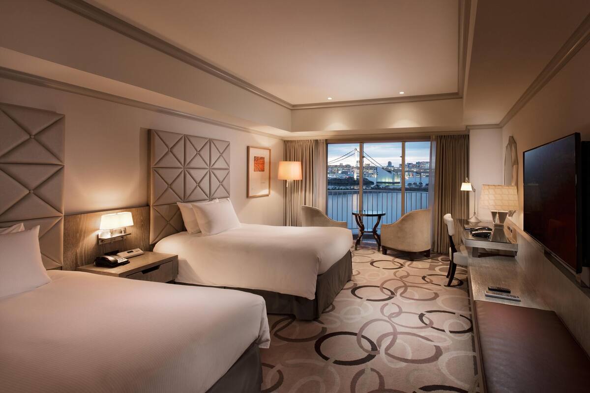 A hotel room with two beds, a desk with a TV, a chair, a view of a bridge and cityscape, and a large window.