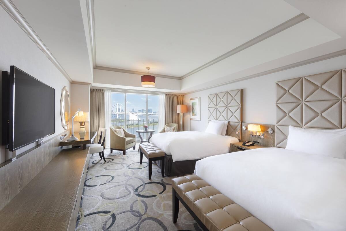 A hotel room with two beds, a TV, a desk, a chair, a lamp, and a view of the city.