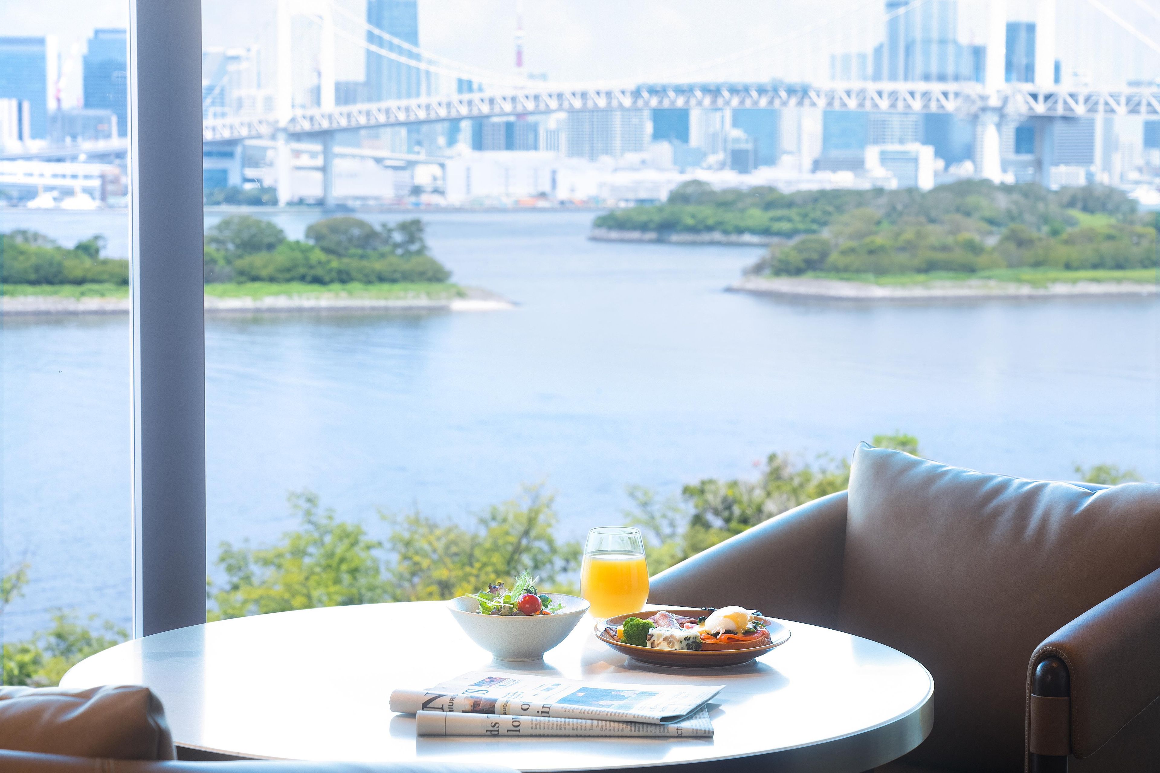 Un comedor con una mesa dispuesta para el desayuno, un periódico y una vista de un río con un puente al fondo.