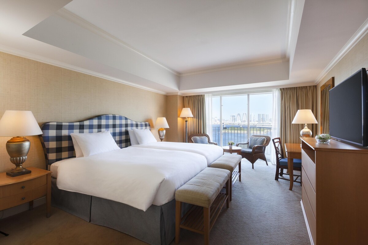 A hotel room with two beds, a desk, a chair, a TV, and a large window with a city view.