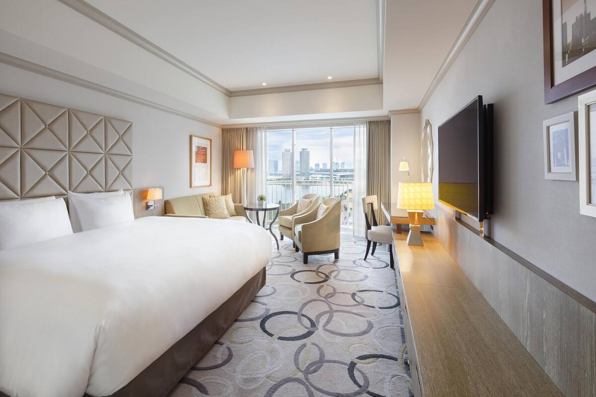 A hotel room with a large bed, a seating area with chairs, a TV, and a city view.