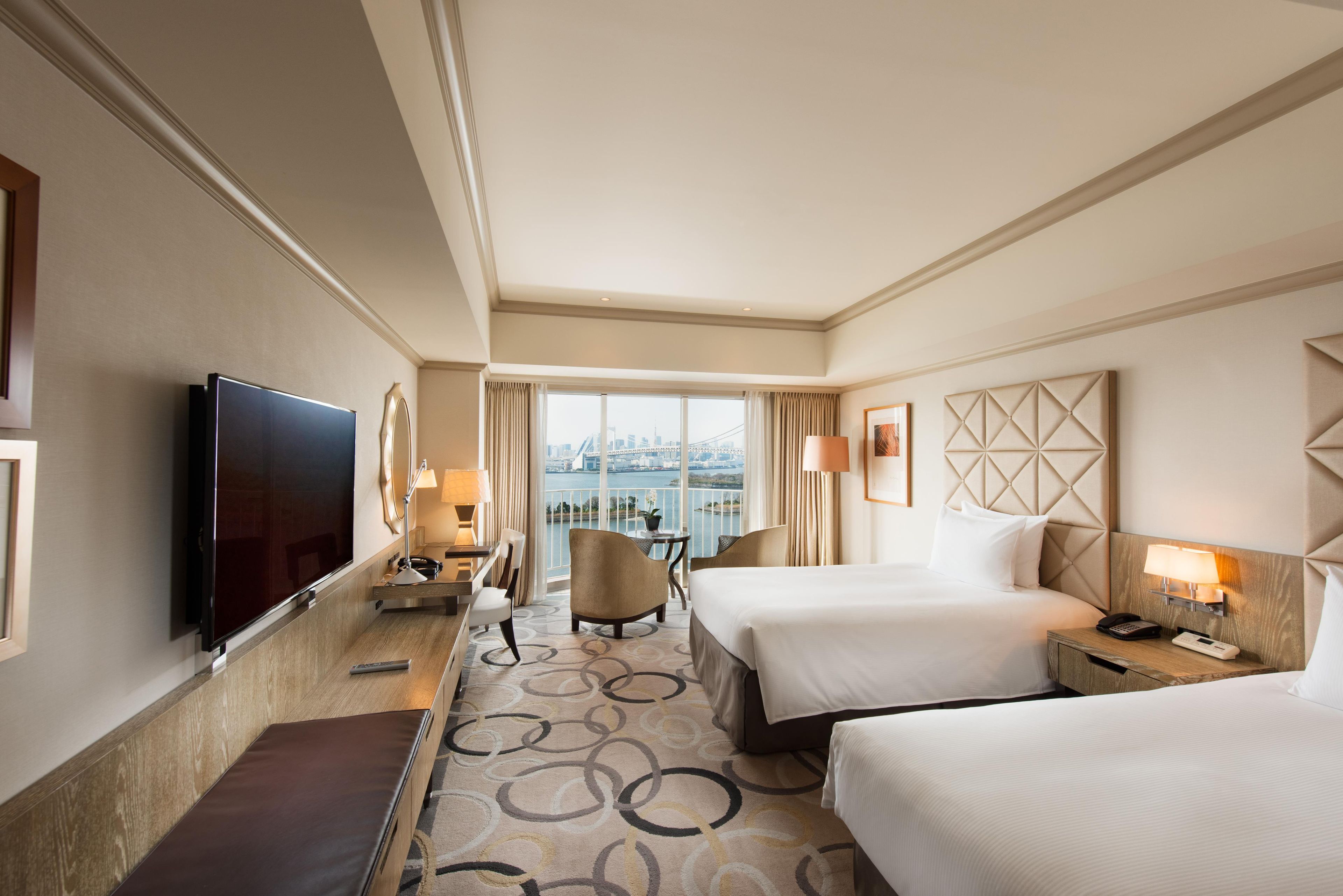 A hotel room with a large bed, a smaller bed, a desk with a lamp, a TV, and a view of a cityscape.