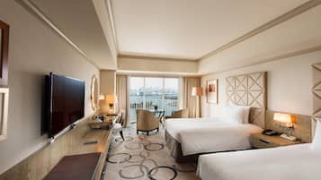 A hotel room with a large bed, a smaller bed, a desk with a lamp, a TV, and a view of a cityscape.