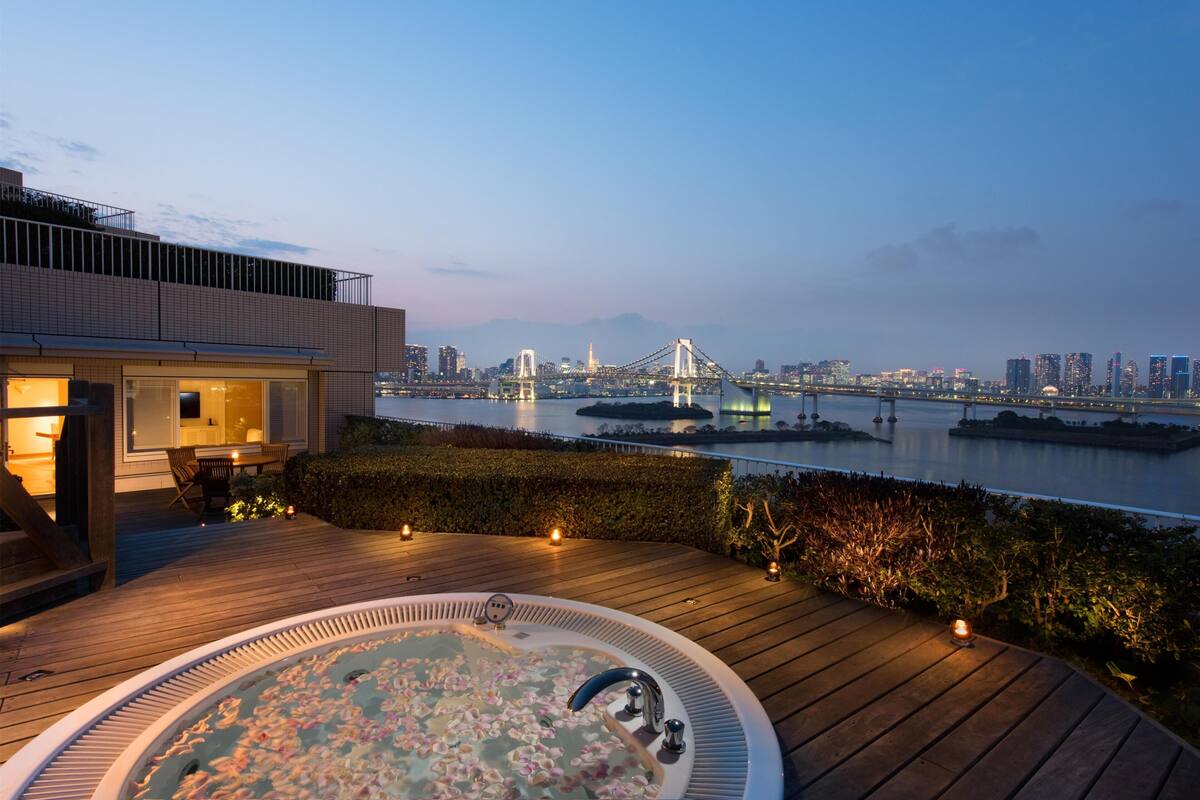 A rooftop terrace with a hot tub, wooden deck, and a cityscape view including a bridge.
