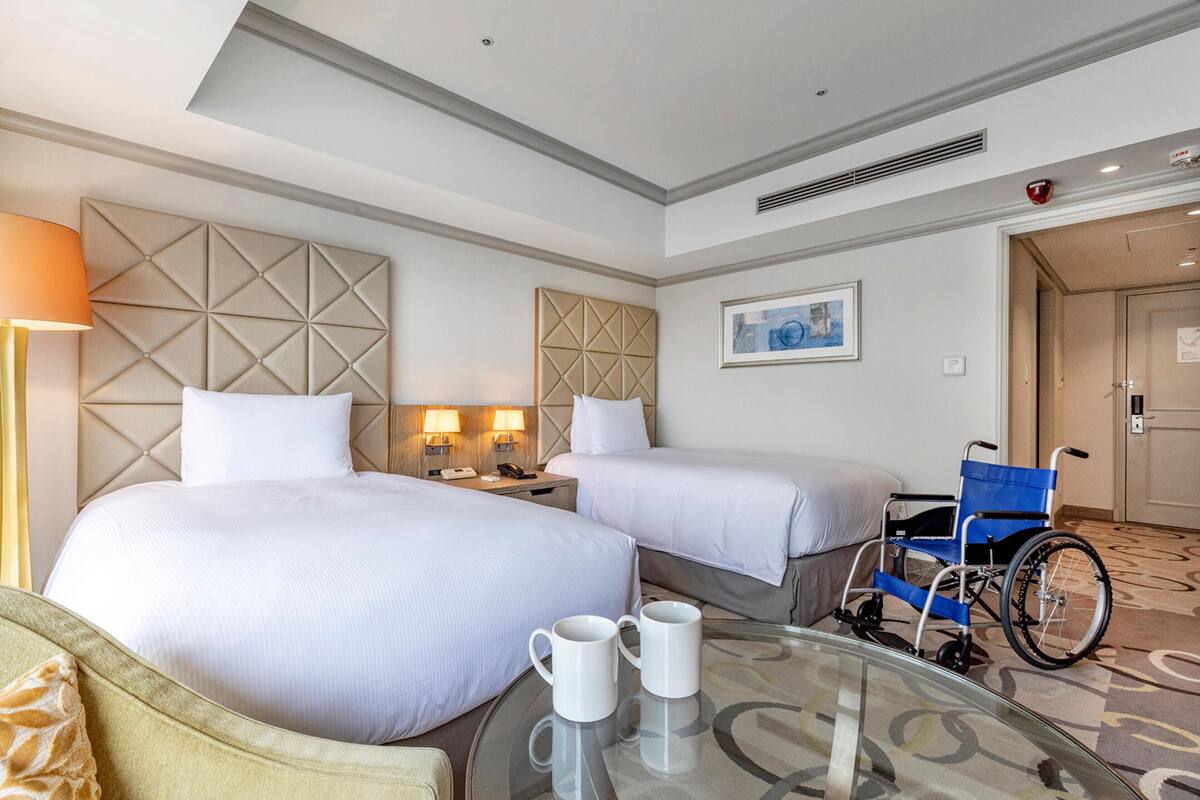 A hotel room with two beds, a wheelchair, a glass table with two mugs, and a painting on the wall.