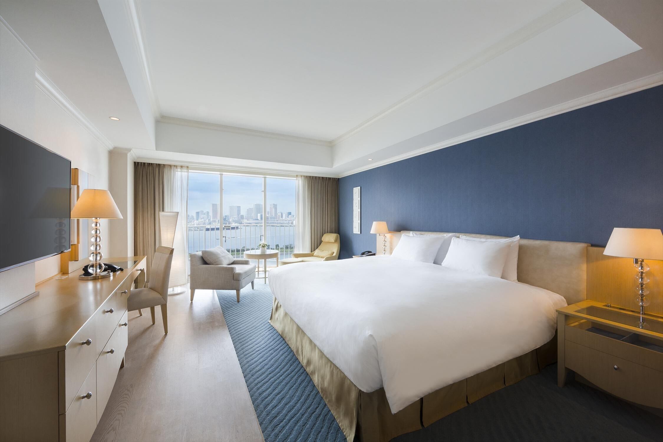 A hotel room with a large bed, a desk, a chair, a small table, and a view of the city.