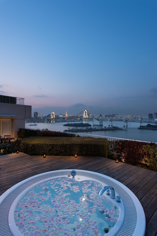 Un bain à remous orné de pétales de rose, installé sur une terrasse en bois offrant une vue sur le paysage urbain et un pont.