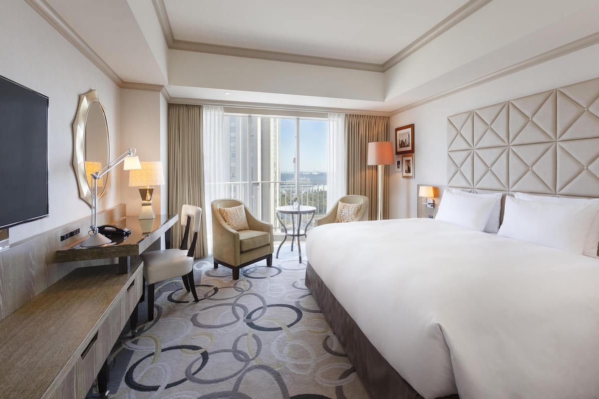 A hotel room with a large bed, a desk with a flat-screen TV, a chair, a small table, and a view of the city.