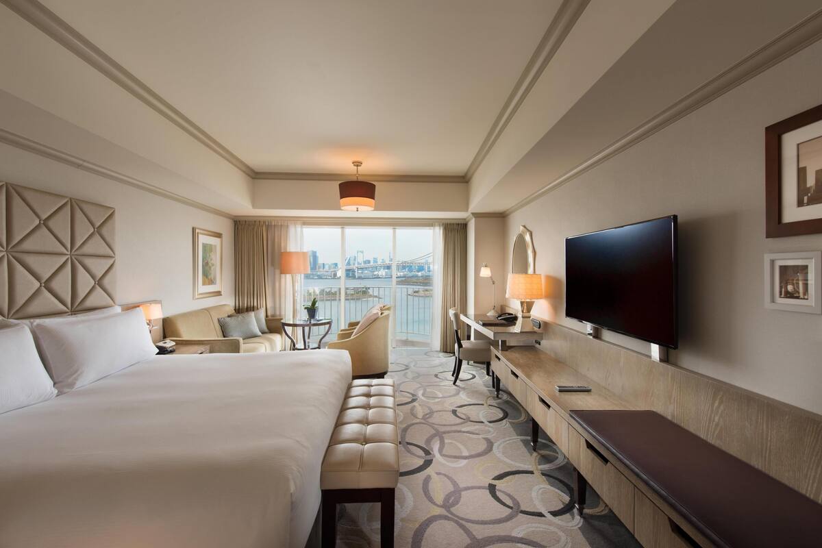 A hotel room with a large bed, a desk, a TV, and a view of a cityscape through a large window.