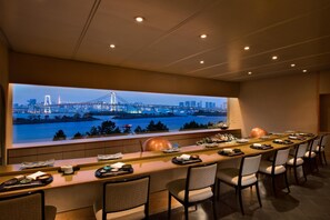 A modern bar area with a long counter, multiple chairs, and a large window with a cityscape view. - Hilton Tokyo Odaiba (Tokyo)