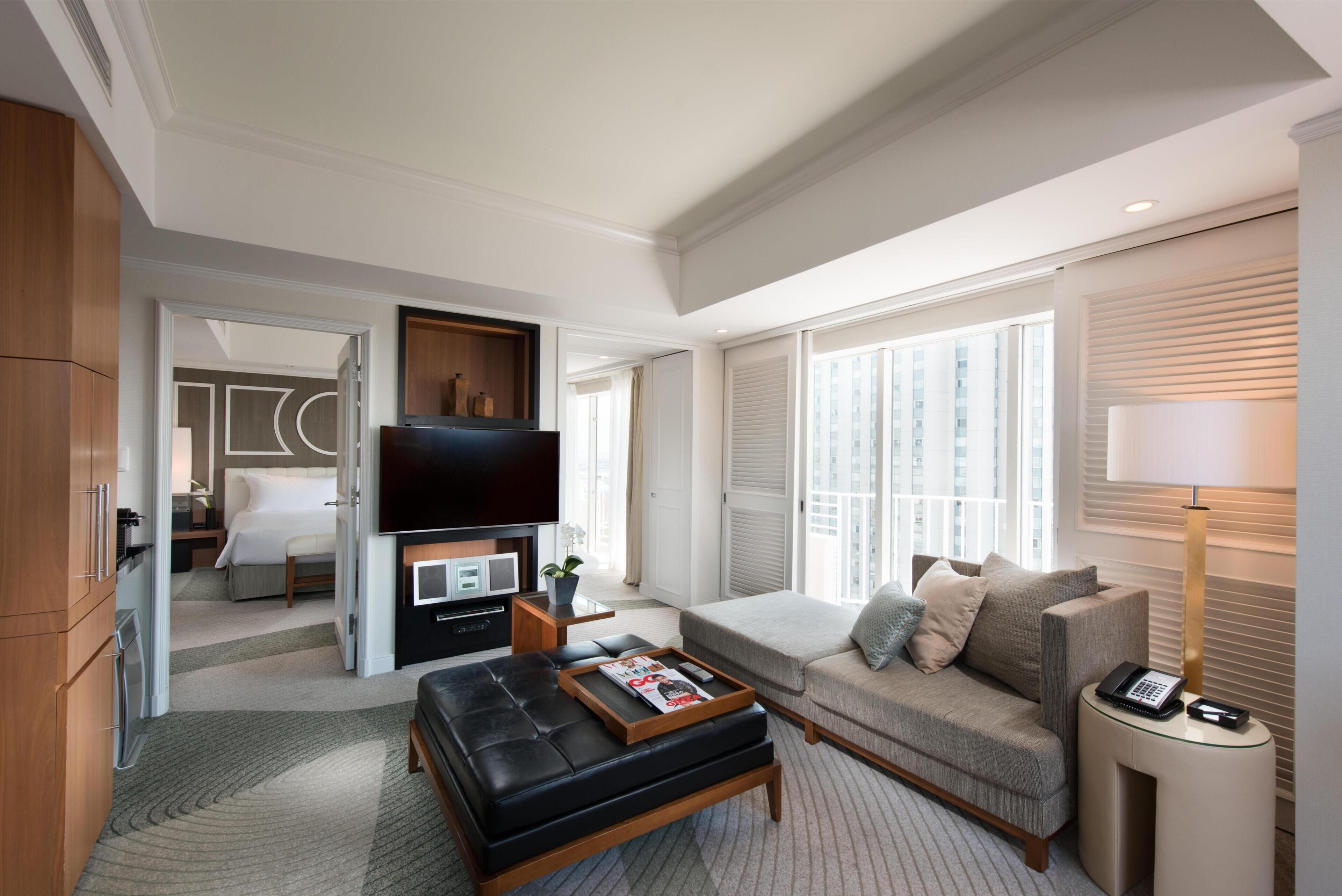 A hotel room with a living area, a sofa, a coffee table, a TV, and a bedside table with a lamp.