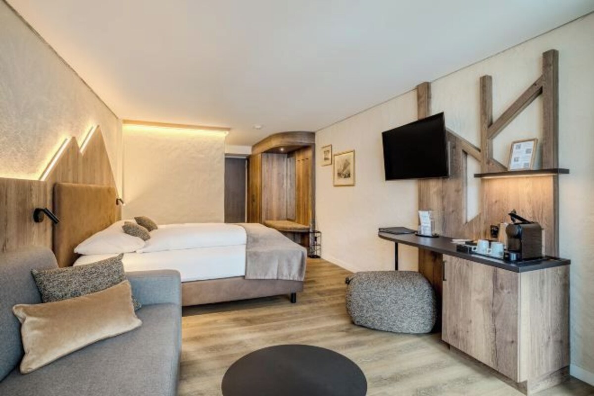 A modern hotel room with a bed, a sofa, a small round table, and a TV mounted on the wall.