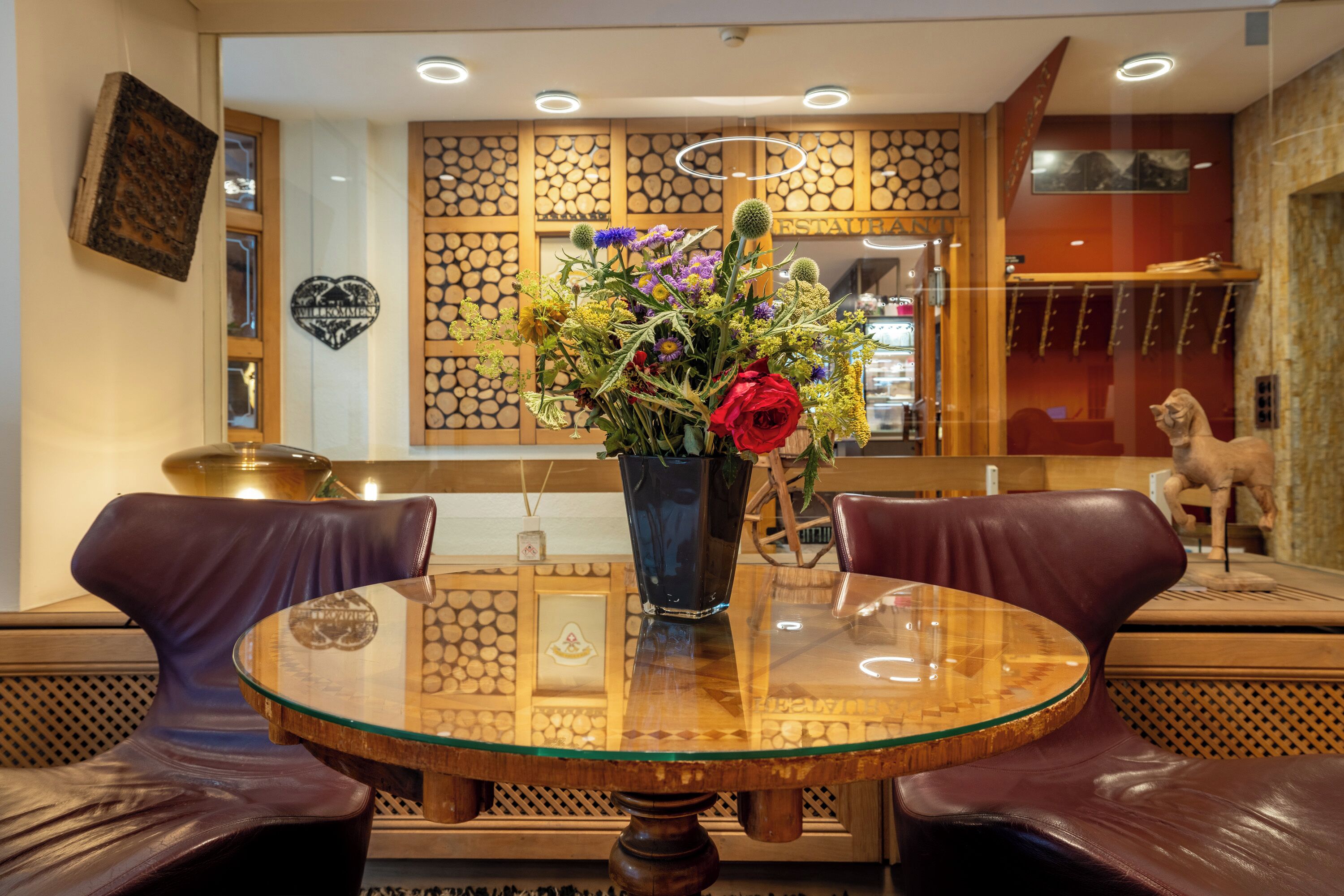 A round glass table with a floral centerpiece, two leather chairs, a decorative wall with a heart-shaped mirror, and a statue on the right.