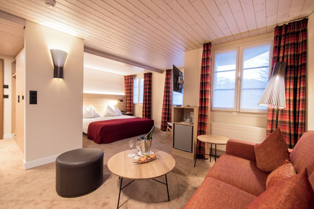 Ein Hotelzimmer mit einem Bett, einem kleinen Tisch mit Flasche und Gläsern, einer Couch, einem Fernseher und einem Fenster mit Vorhängen.