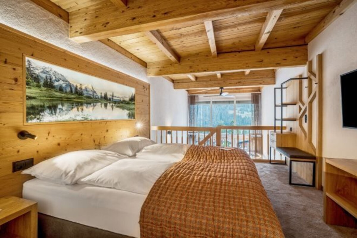 A bedroom with a wooden ceiling, a bed with white bedding, a painting of a mountain landscape, and a balcony with a view.