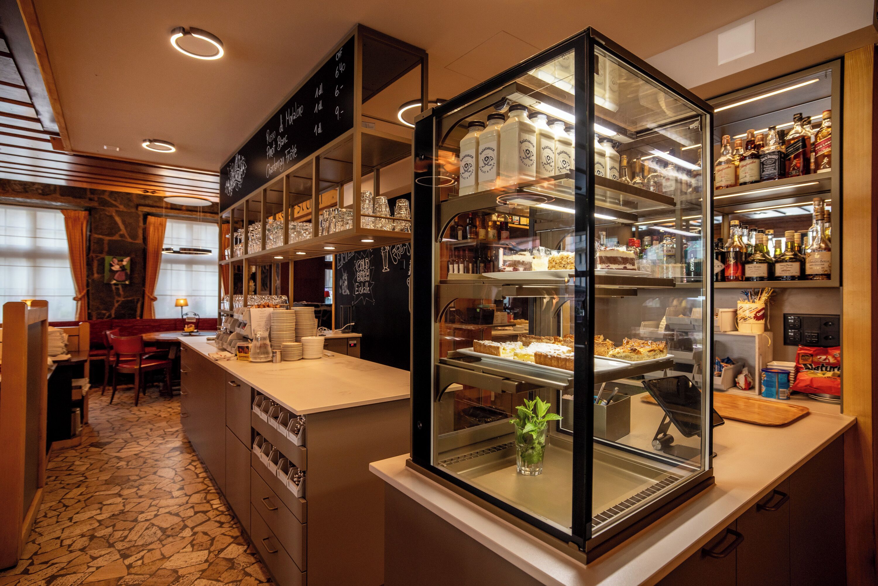 A bakery counter with a glass display case, shelves with baked goods, and a chalkboard menu.