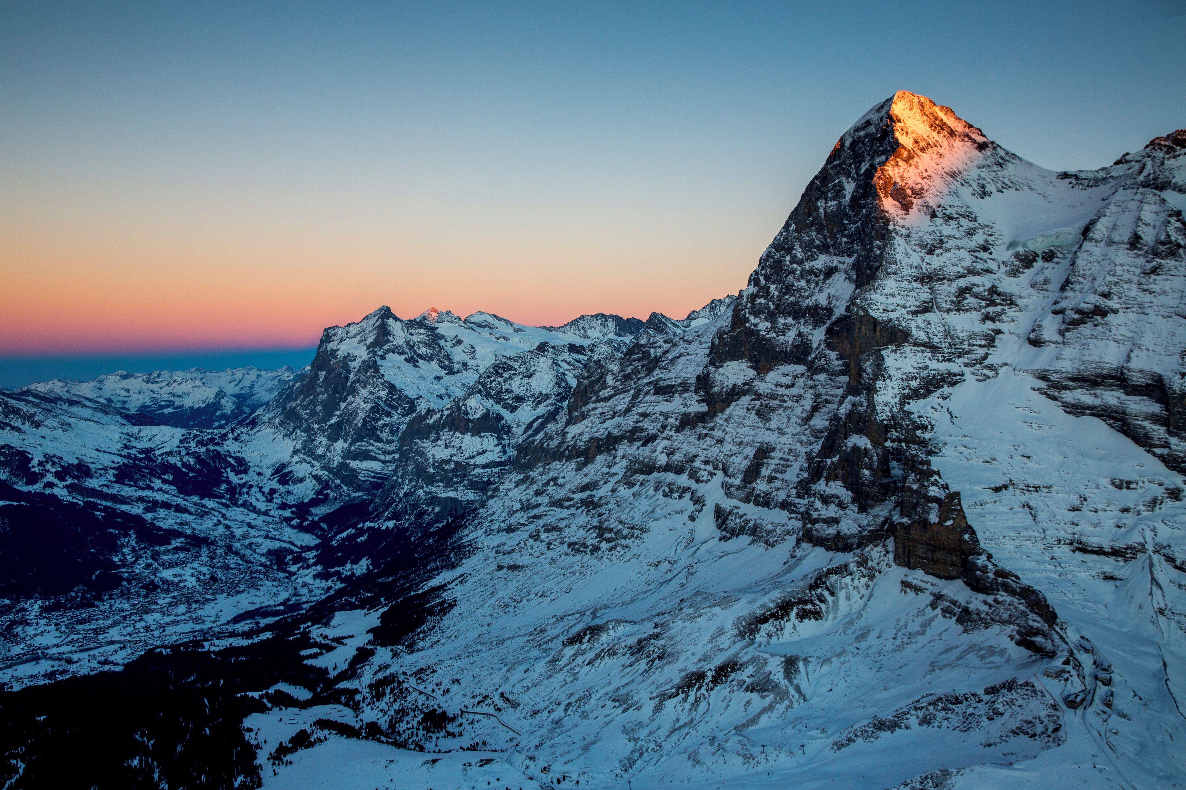 A snow-covered mountain peak at sunset with rugged terrain and rocky outcrops.