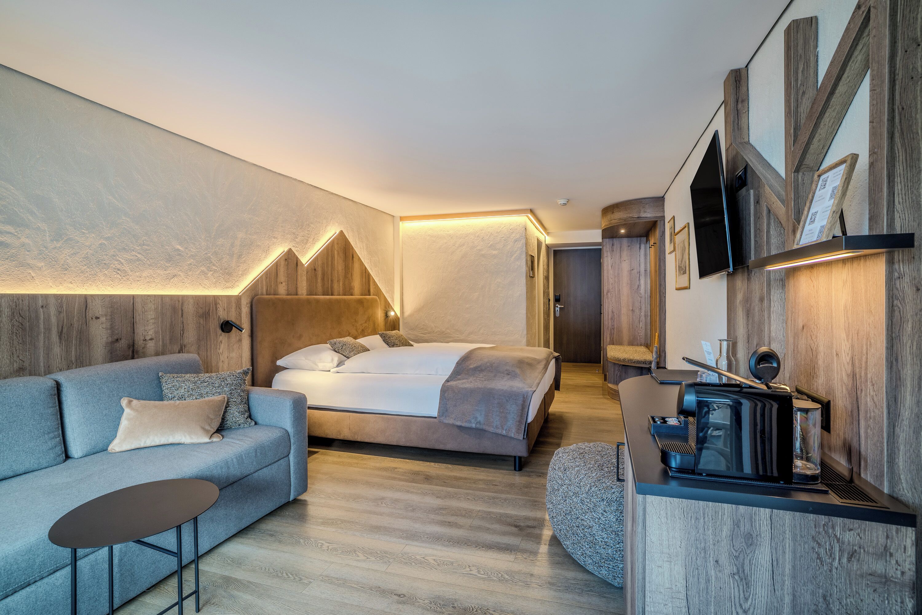 A modern hotel room with a bed, sofa, coffee table, and a small kitchenette area.
