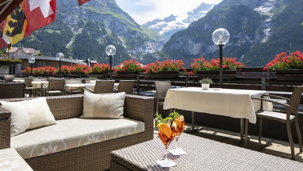 An outdoor seating area with wicker furniture, tables with white tablecloths, and a mountain view. - Hotel Kreuz & Post (Grindelwald)