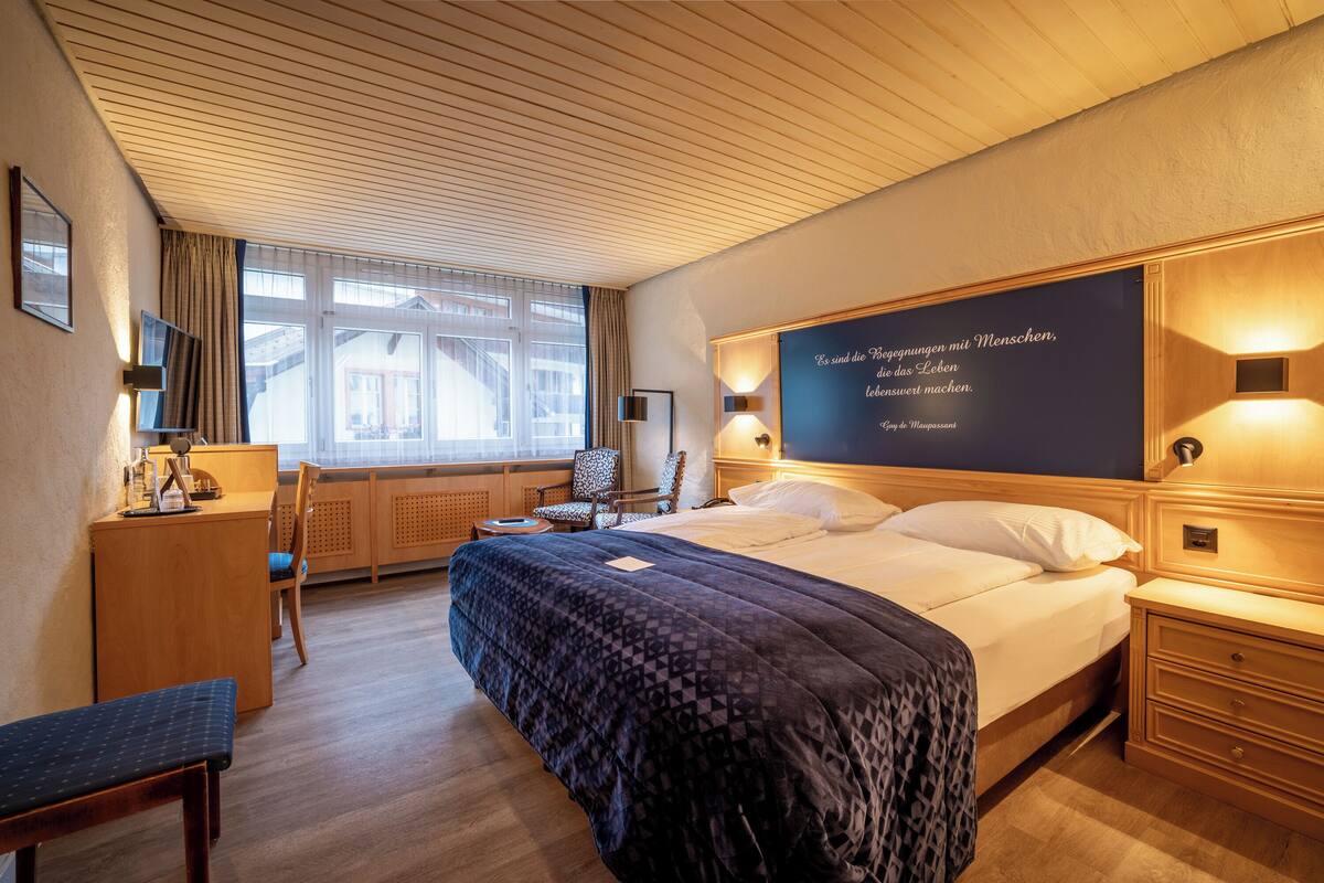 Ein Hotelzimmer mit einem großen Bett, einem Schreibtisch, einem Stuhl und einem fensterbepflanzten Fenster.