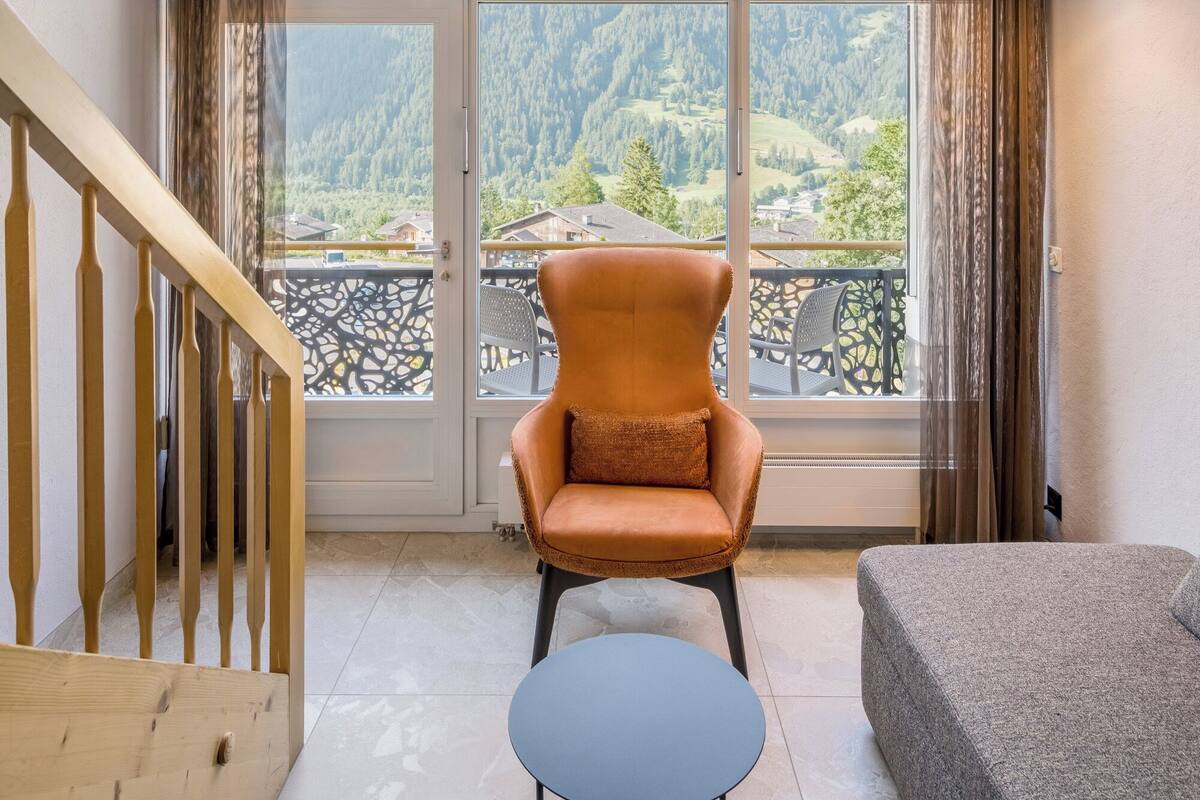 Ein Zimmer mit großem Fenster und Blick auf die Berge, einer Holztreppe, einem braunen Sessel und einem grauen Sofa.