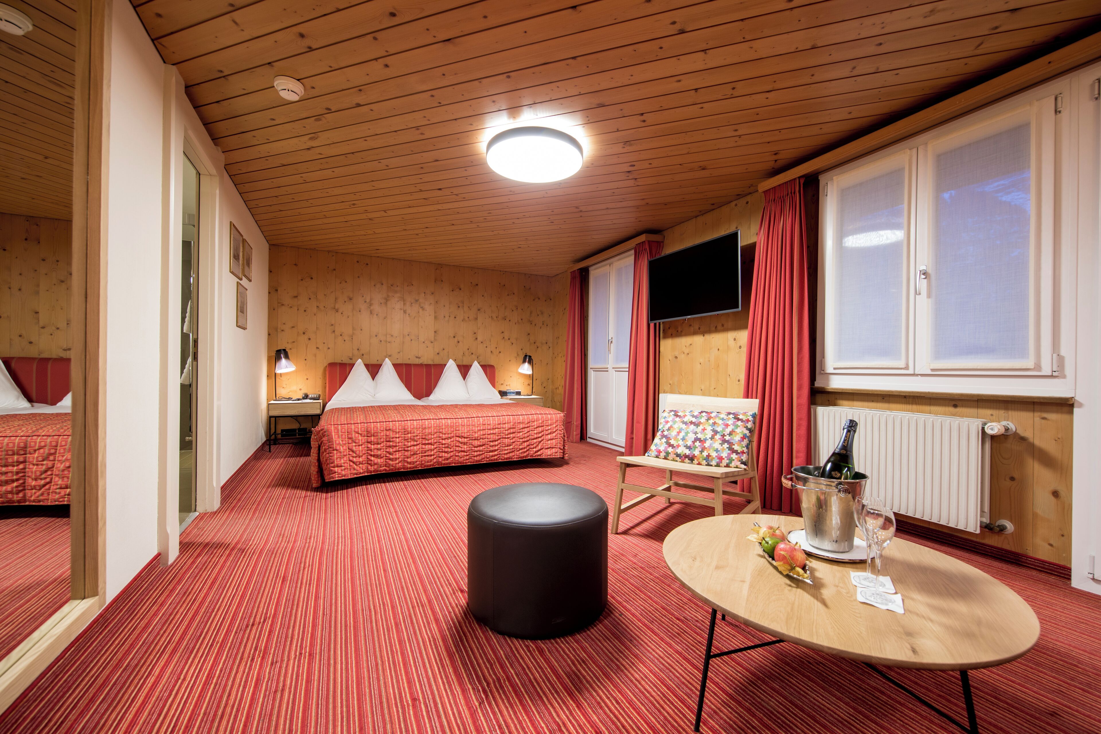Una habitación de hotel con techo de madera, dos camas con ropa de cama roja, una mesita con una botella y un vaso, y un radiador.
