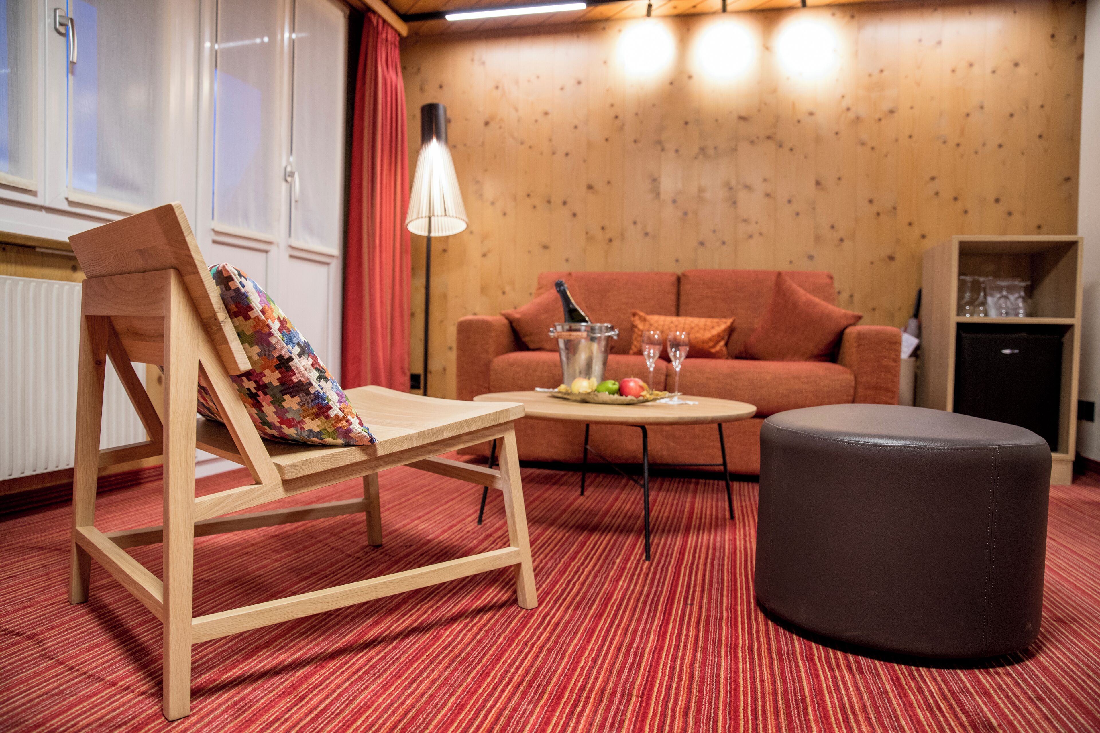 Ein gemütlicher Raum mit Holzboden, einem rot gestreiften Teppich, einem Holzstuhl mit farbigem Kissen, einem kleinen runden Tisch mit Flasche und Gläsern, einem braunen Hocker, einer roten Couch und einem Holzschrank.