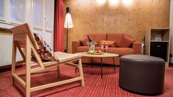 Ein gemütlicher Raum mit Holzboden, einem rot gestreiften Teppich, einem Holzstuhl mit farbigem Kissen, einem kleinen runden Tisch mit Flasche und Gläsern, einem braunen Hocker, einer roten Couch und einem Holzschrank.