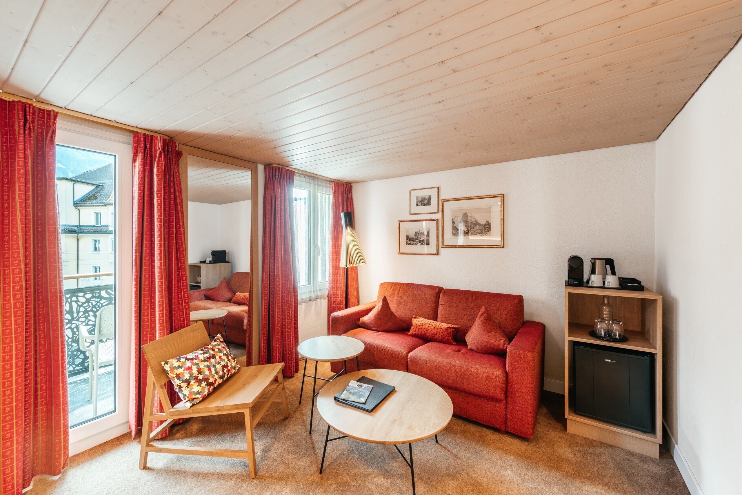 Una sala de estar acogedora con un sofá rojo, una mesa de centro de madera y una mesita auxiliar. Hay una estantería con adornos y un mueble. La habitación tiene ventanas grandes con cortinas rojas y vistas al exterior.
