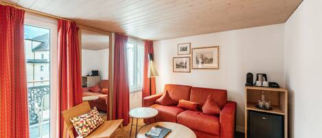 Una sala de estar acogedora con un sofá rojo, una mesa de centro de madera y una mesita auxiliar. Hay una estantería con adornos y un mueble. La habitación tiene ventanas grandes con cortinas rojas y vistas al exterior.