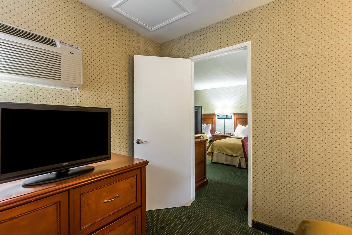 A hotel room with a television on a wooden dresser, a white door leading to another room, and patterned wallpaper.