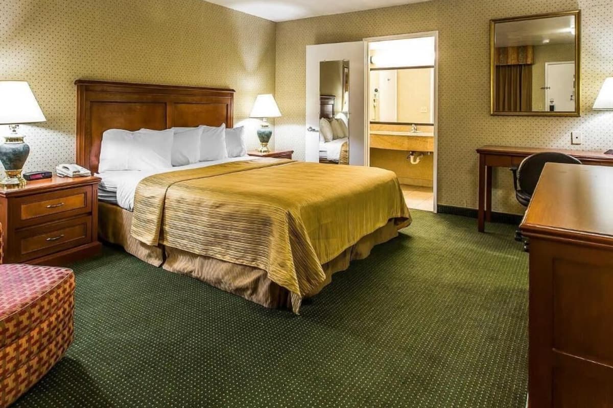 A hotel room with a large bed, bedside tables, a desk, a chair, a mirror, and a bathroom visible through an open door.