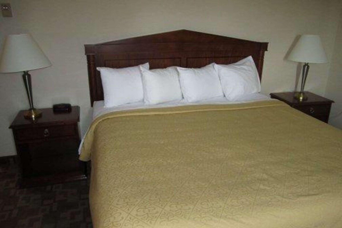 Una habitación de hotel estándar con cabecera de madera, cama con ropa blanca, colcha beige estampada, dos mesitas de noche con lámparas y una pequeña mesita auxiliar.