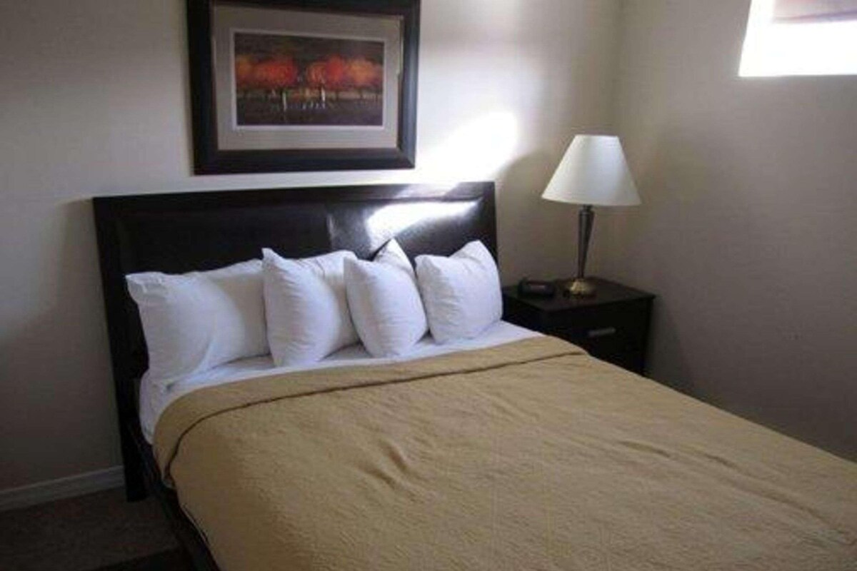 Una cama bien tendida con ropa blanca y un plaid color beige, un cuadro en la pared y una l ámpara de noche.