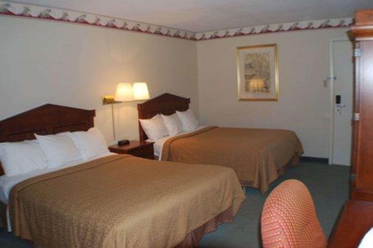 Habitación de hotel con dos camas, una mesita de noche con lámpara, un cuadro en la pared y una puerta.