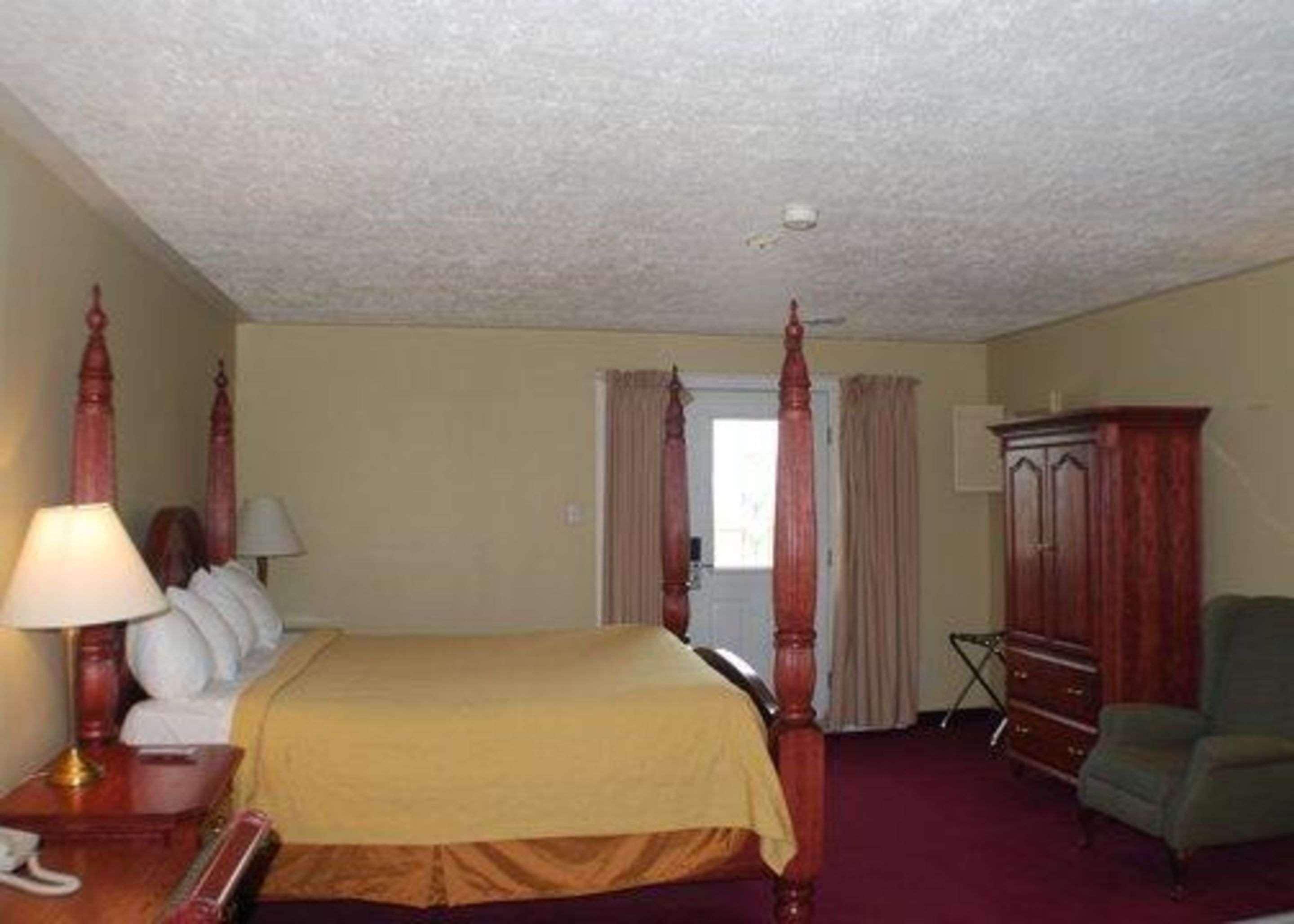 A four-poster bed with a yellow bedspread, a wooden nightstand with a lamp, a green armchair, and a wooden cabinet.