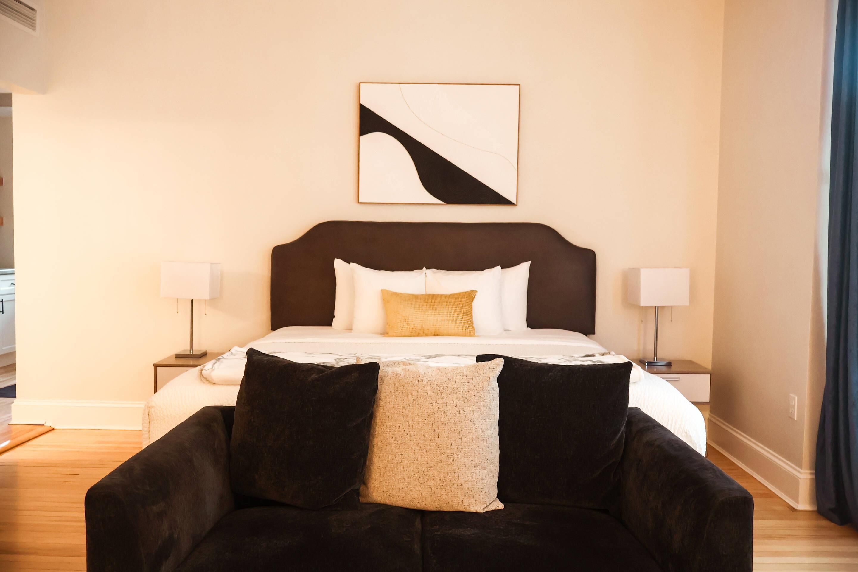A hotel room with a bed, two bedside tables with lamps, a dark-colored sofa with cushions, and a wall-mounted abstract art piece.