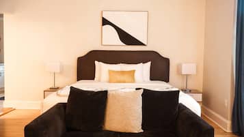 A hotel room with a bed, two bedside tables with lamps, a dark-colored sofa with cushions, and a wall-mounted abstract art piece.